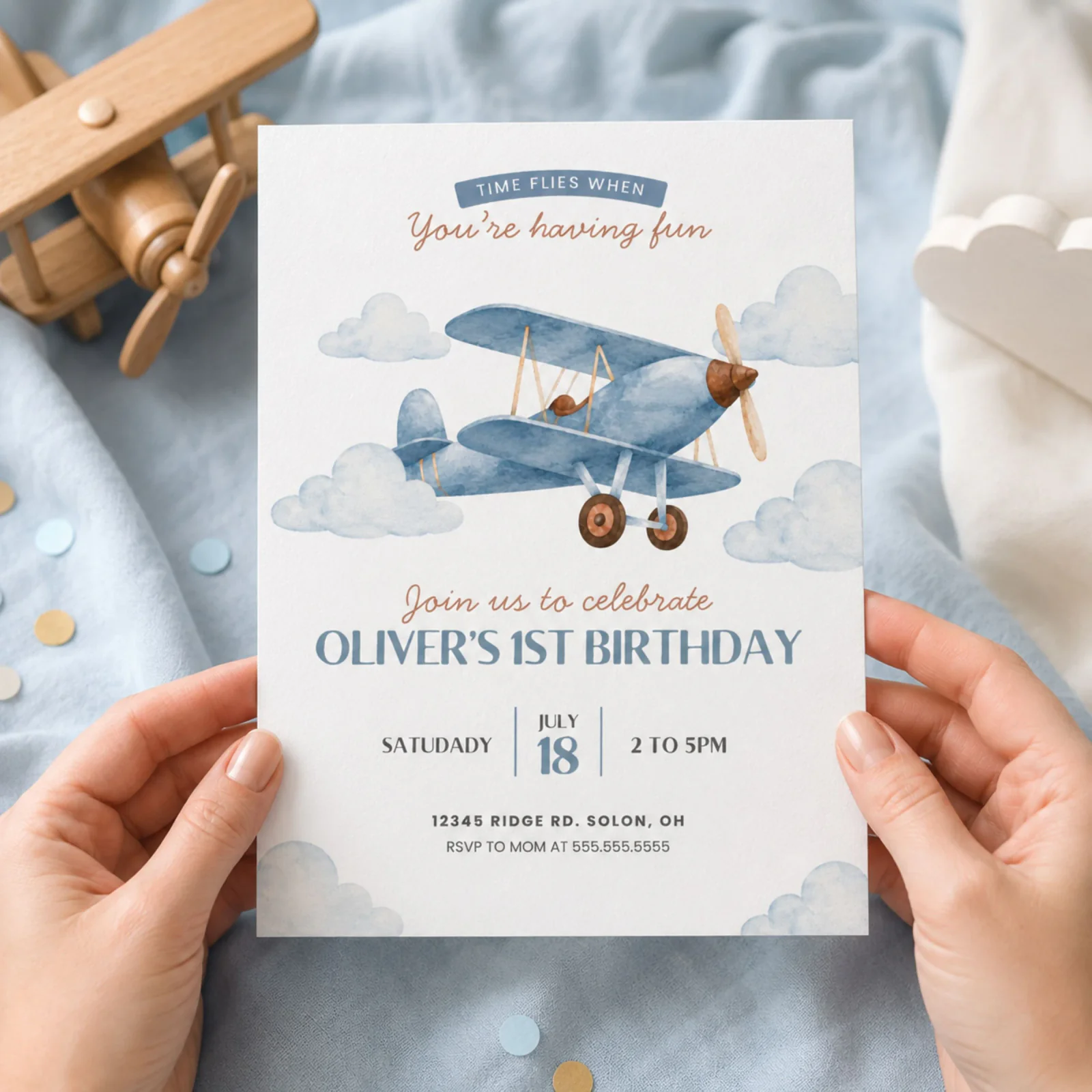 Aviation 1st birthday invitation held in hands, featuring a blue watercolor airplane and clouds.