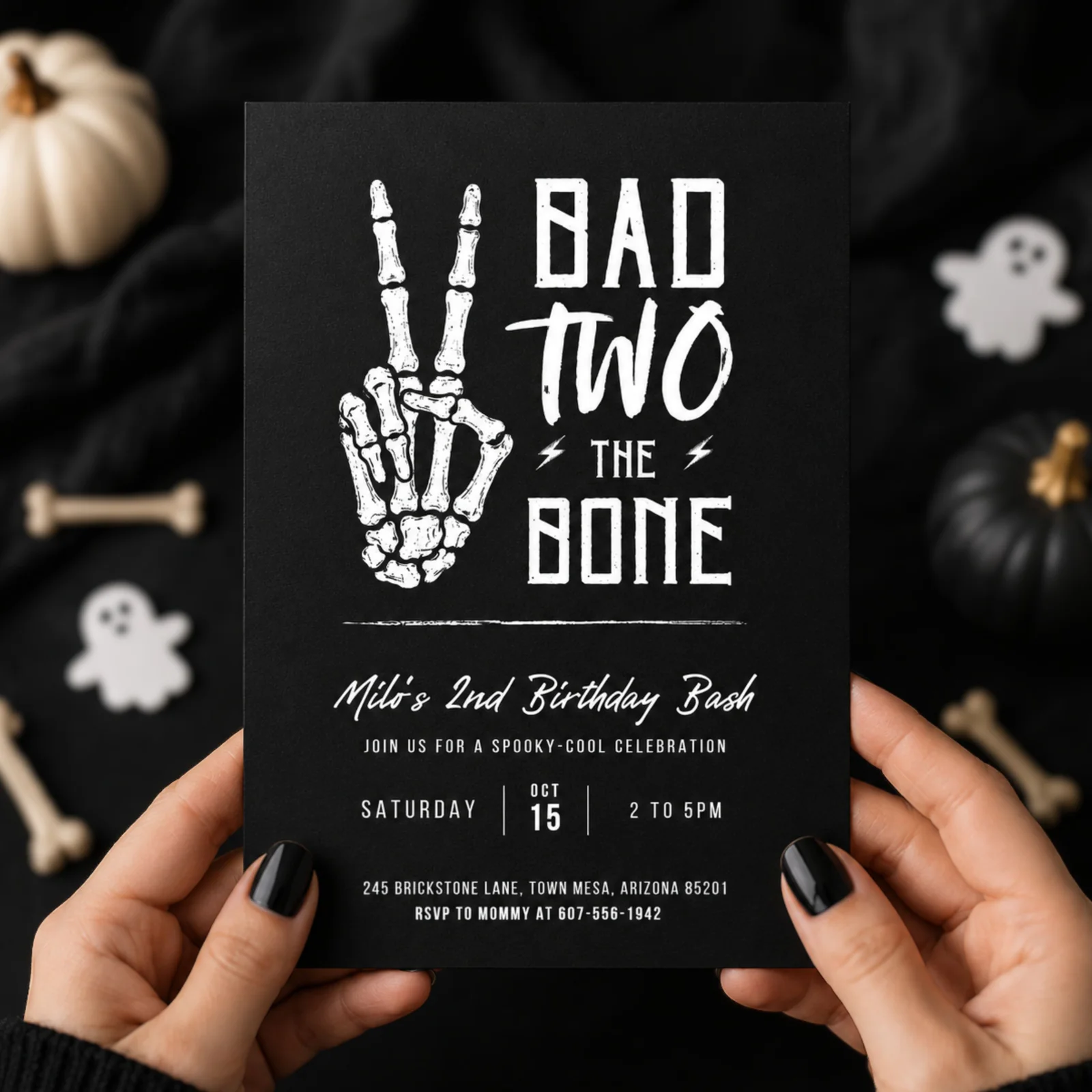 Bad Two the Bone birthday invitation held in hands, featuring a skeleton peace sign on a black background, an editable Canva template.