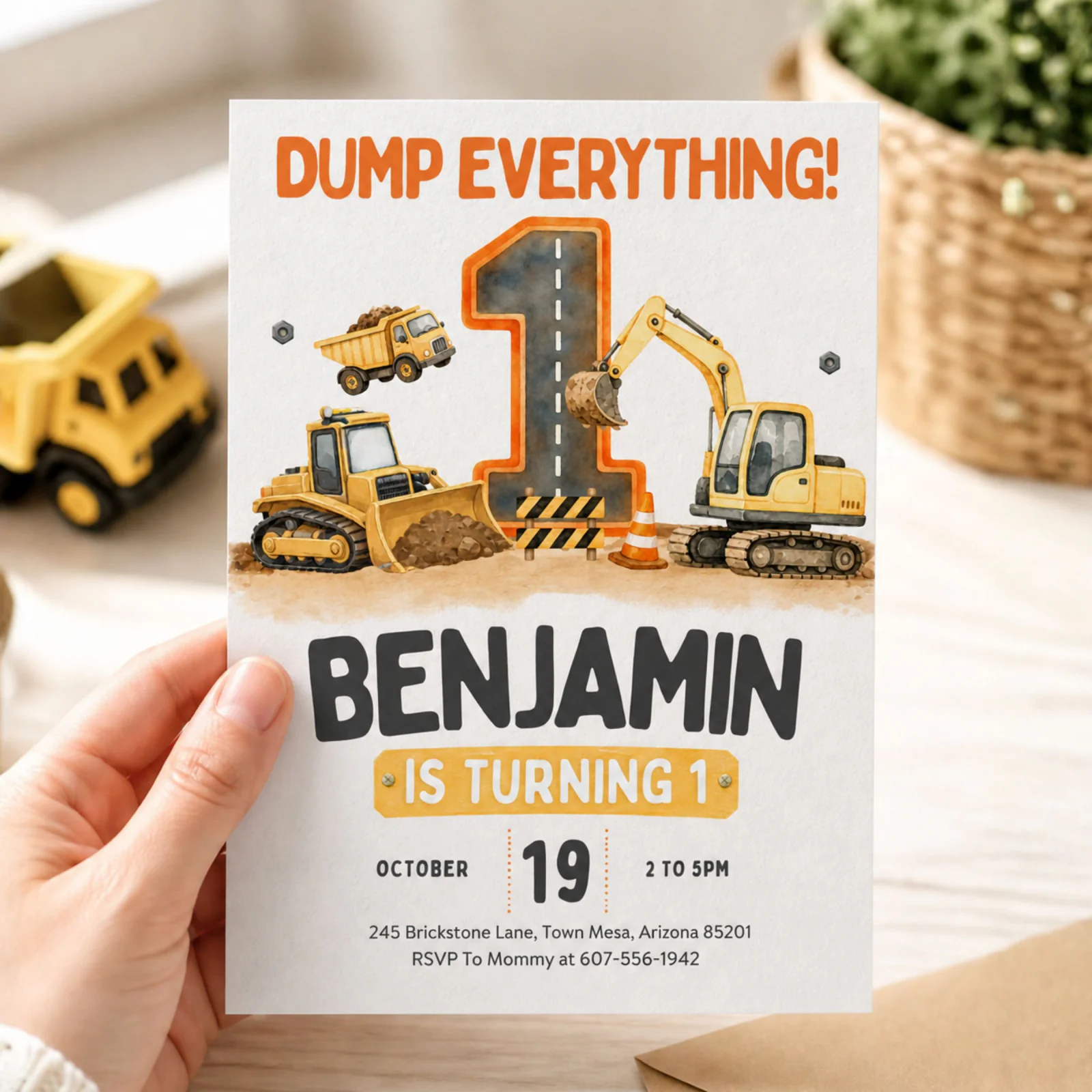 Construction 1st birthday invitation held in hand, featuring a dump truck and excavator design, an editable Canva template.