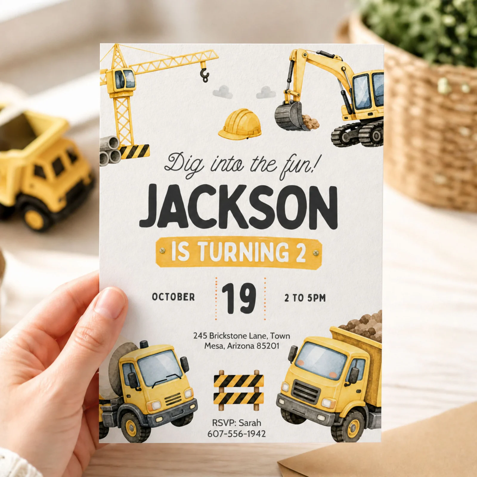 Construction birthday invitation held in hand, featuring a yellow dump truck and crane design.