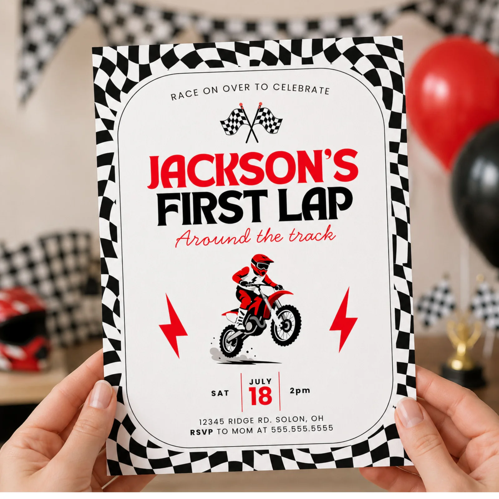Dirt bike 1st birthday invitation held in hands, featuring the red motocross rider design with checkered flag border.