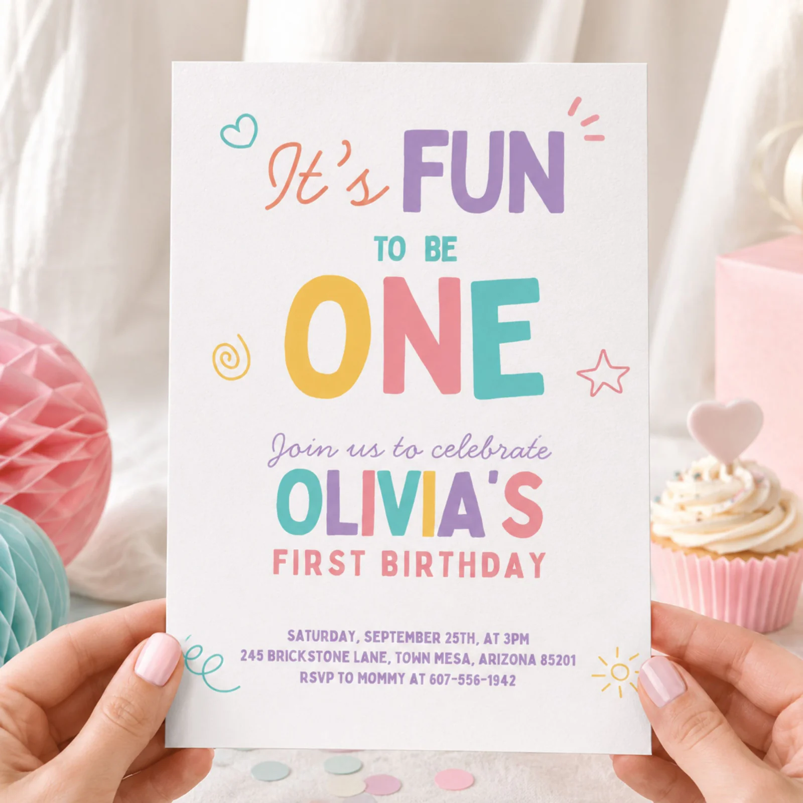 Pastel girl first birthday invite printed invitation card held in hands, featuring colorful 'It's Fun To Be One' text and playful doodles.