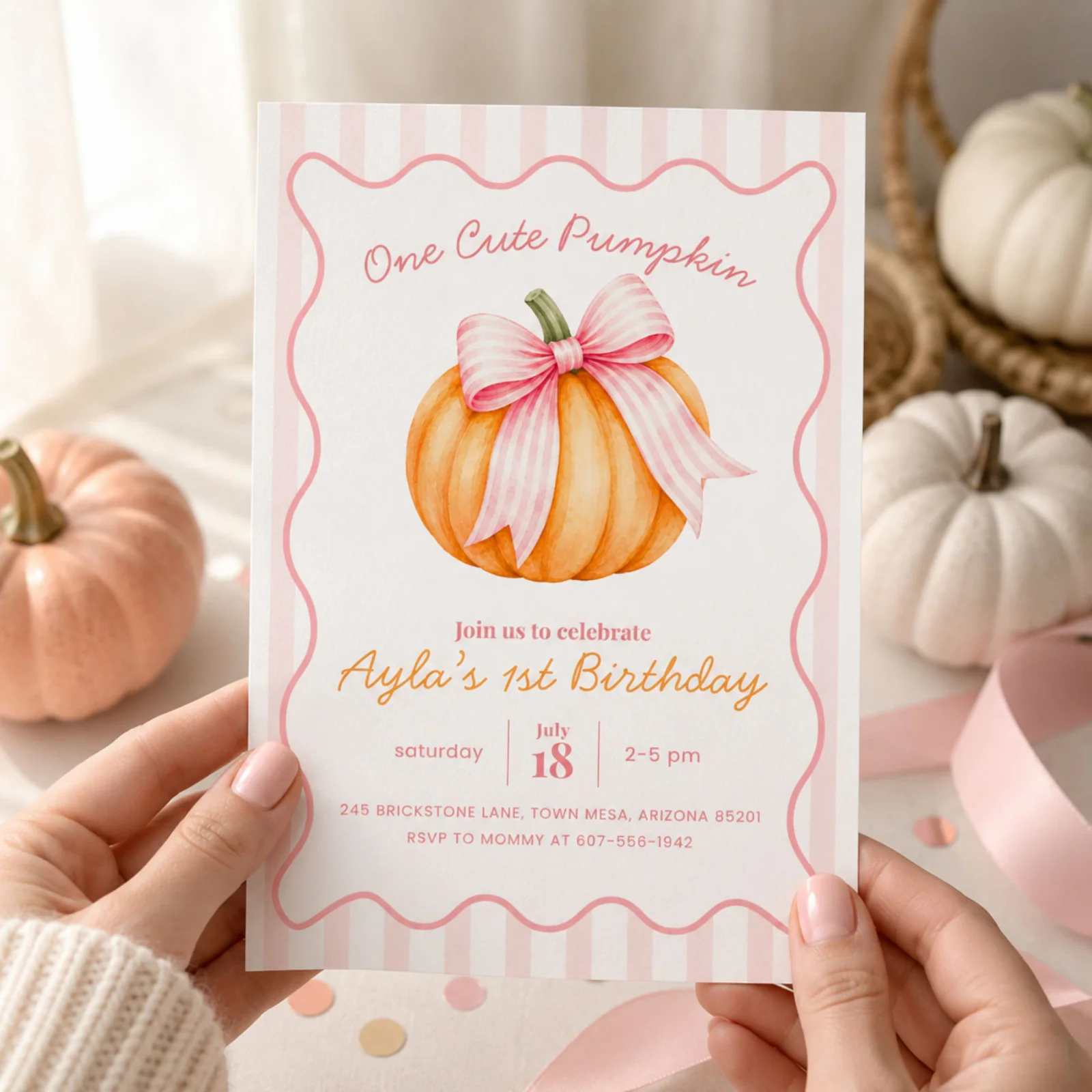 Pumpkin First Birthday Invitation held in hands, featuring a pink striped bow pumpkin on a blush background, an editable Canva template.