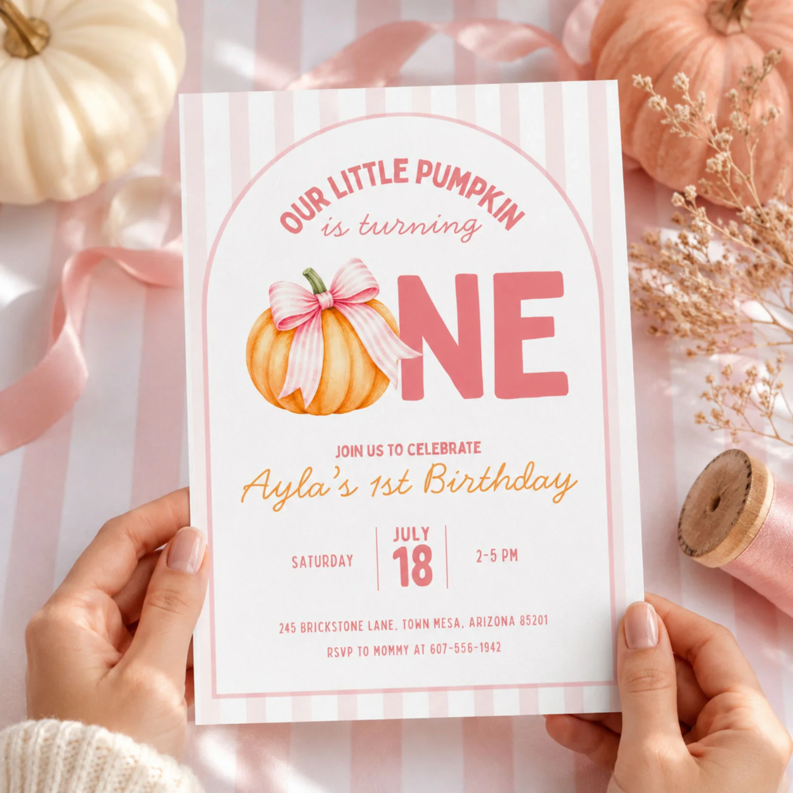 Pumpkin first birthday invitation held in hands, showcasing the girly pumpkin and pink striped bow design. Editable Canva template.