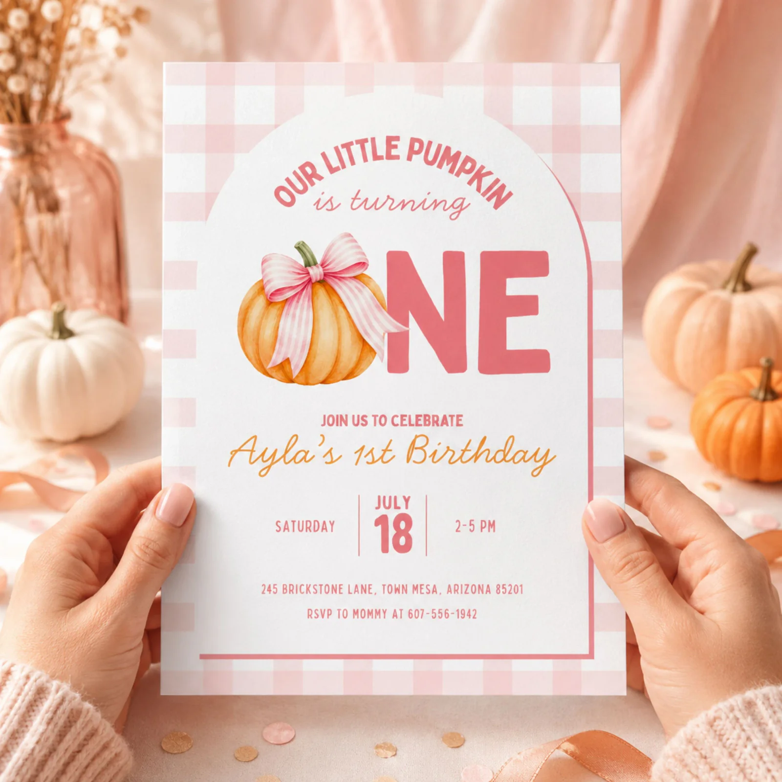 Pumpkin First Birthday Invitation held in hands, featuring a pink gingham background and a watercolor pumpkin with a pink bow, an editable Canva template.